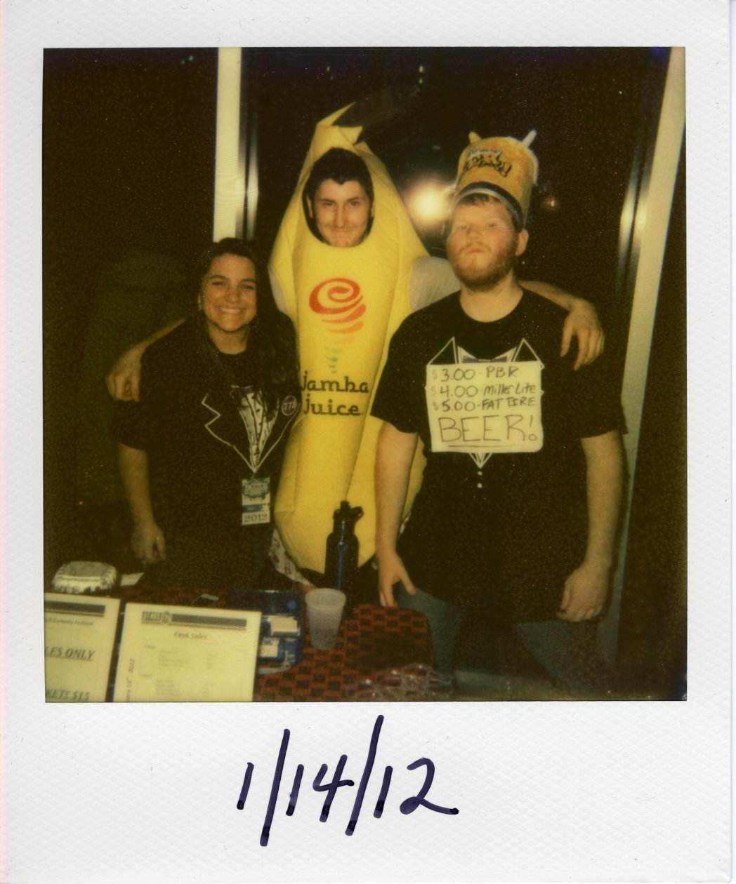 Three people standing at table. From left to right, a woman wearing a t-shirt printed to look like a fake tuxedo, a man wearing a banana costume with a Jamba Juice logo printed on it, and a man wearing a sign advertising beer prices: $3.00 - PBR $4.00 Miller Lite $5.00 Fat Tire BEER!