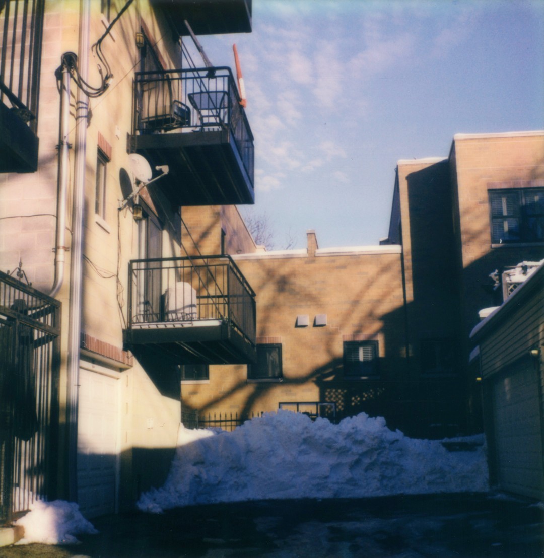 An alleyway in Chicago, on the left we can see a row of balconies, on the right we can see some garages and in the center there is a plowed drive way ending in a large snowpile.